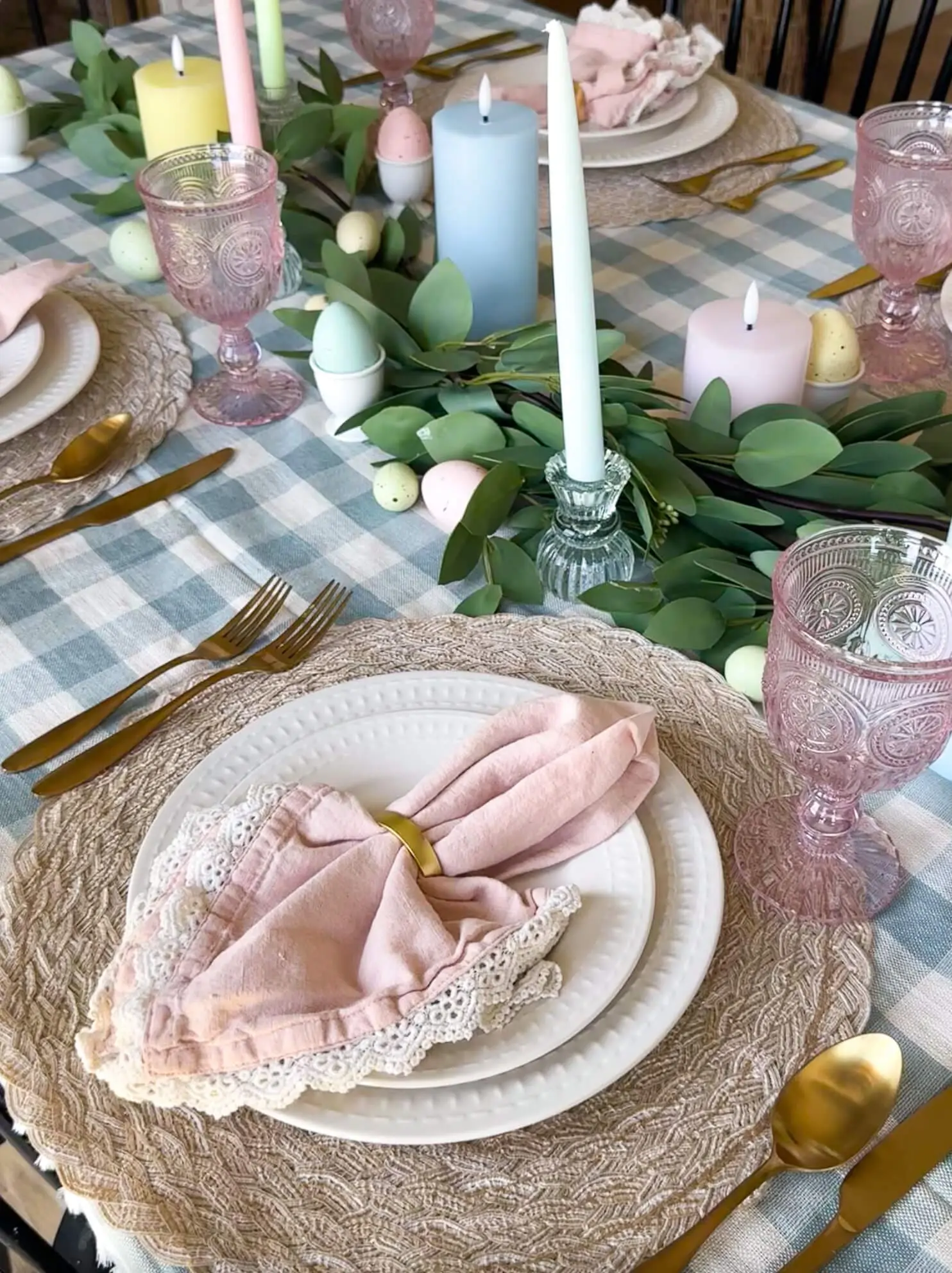 Close-up of Easter table decor featuring blush lace-trimmed napkins, layered white plates, gold flatware, and pastel candles surrounded by eucalyptus and speckled eggs.